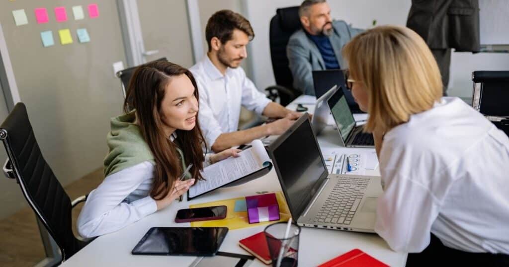 wo people discuss documents at a desk with laptops and tablets, while another person works on a laptop in the background.